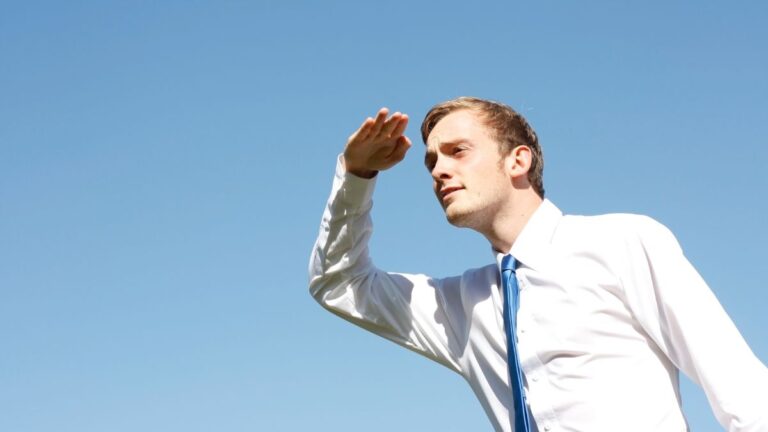 Mężczyzna w białej koszuli i krawacie patrzy w dal, osłaniając oczy dłonią, symbolizując poszukiwanie lub analizę przyszłości na tle błękitnego nieba.