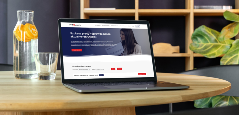 Laptop na drewnianym stole z otwartą stroną internetową HRQuality, prezentującą aktualne oferty pracy w branży technicznej dla osób planujących zmianę branży bez zaczynania kariery od zera. Obok laptopa karafka z wodą z pomarańczą oraz szklanka, w tle roślina i drewniany regał.
